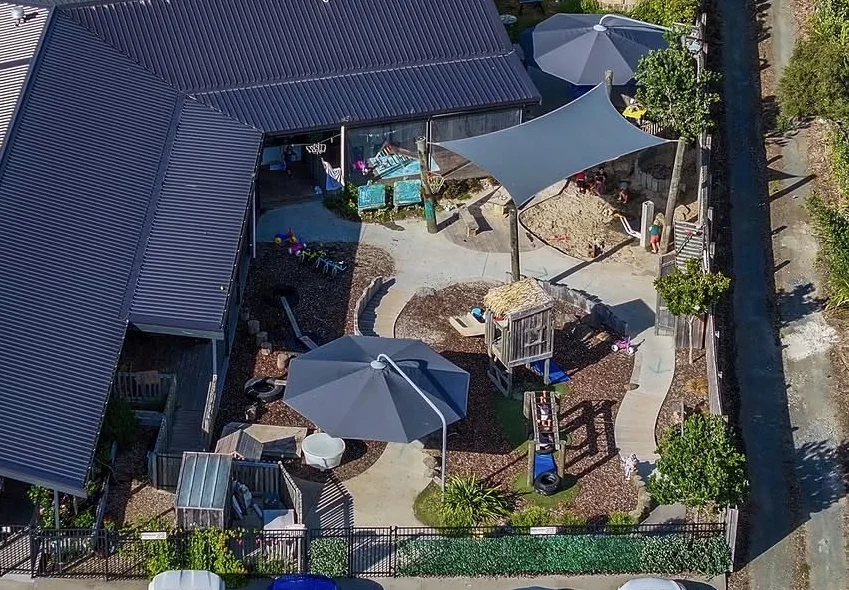 The centre from above: the building wrapping around a courtyard, with shade sails, a sandpit, the fort and planted garden beds.