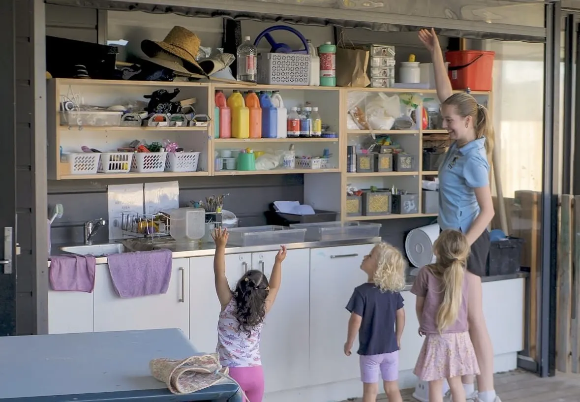 The covered art area with its doors folded open to the courtyard, a teacher and three children reaching up to the paint shelves.