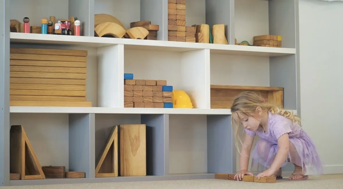 A wall of open shelving filled with wooden blocks, ramps and arches at child height, with a child crouched on the floor building beside it.