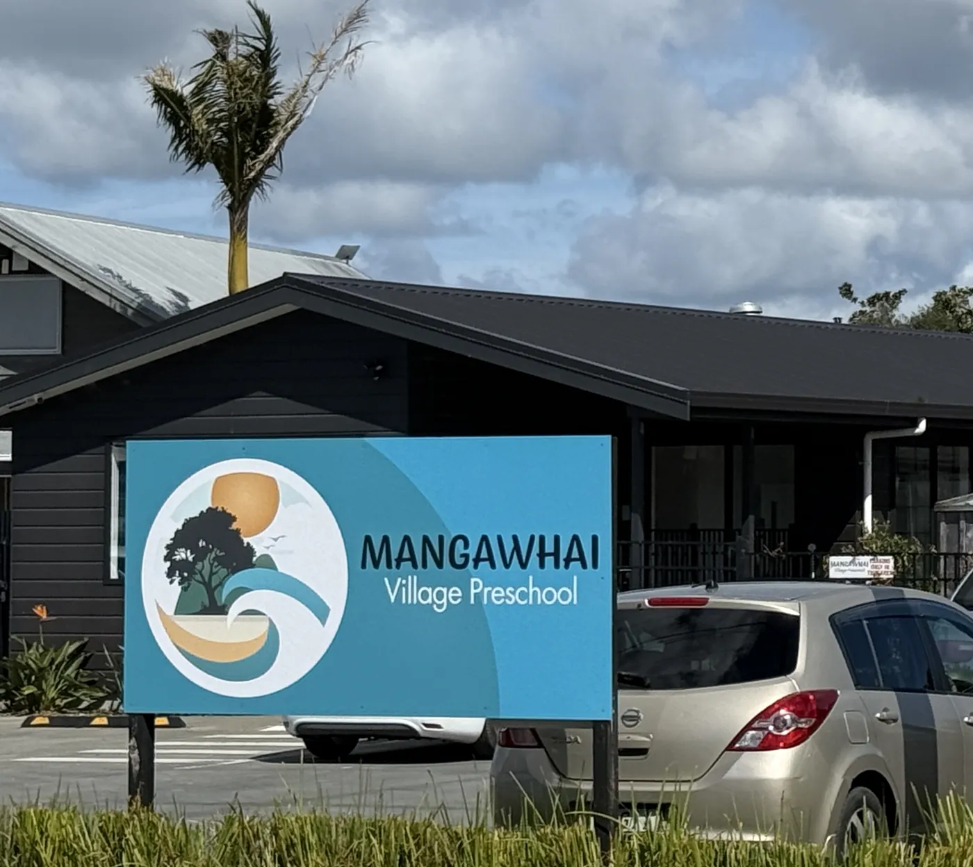 The preschool seen from Molesworth Drive: a low dark timber building behind its sign showing the Mangawhai Village Preschool logo, with a nikau palm beside it.