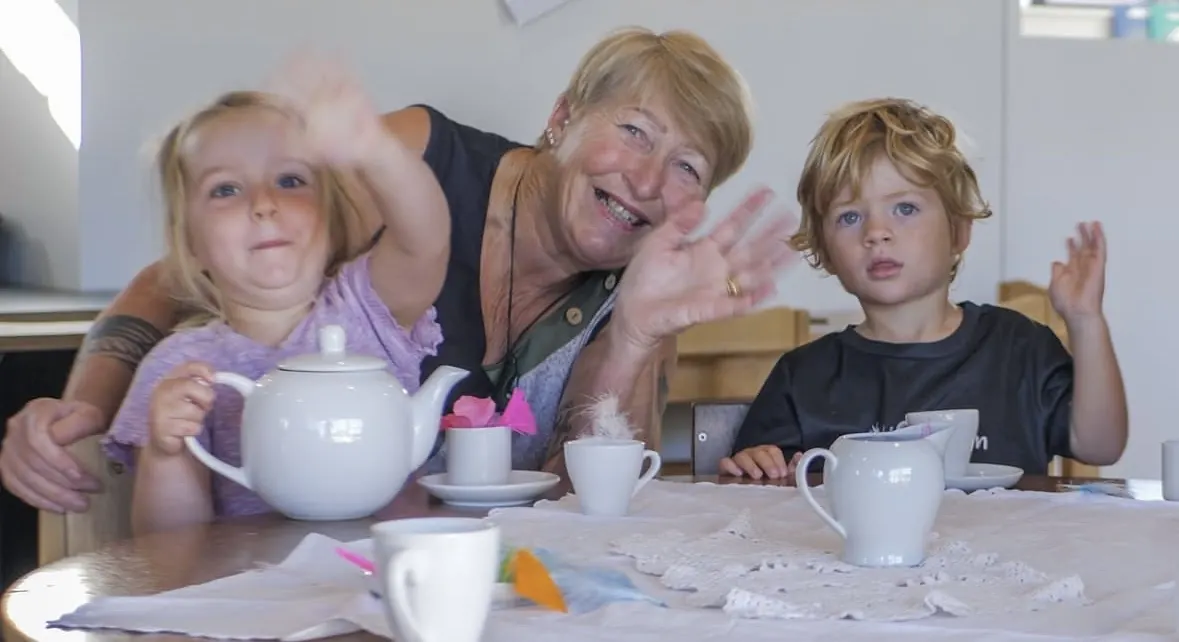 A teacher and two children at a table set with a teapot and cups, all waving at the camera.