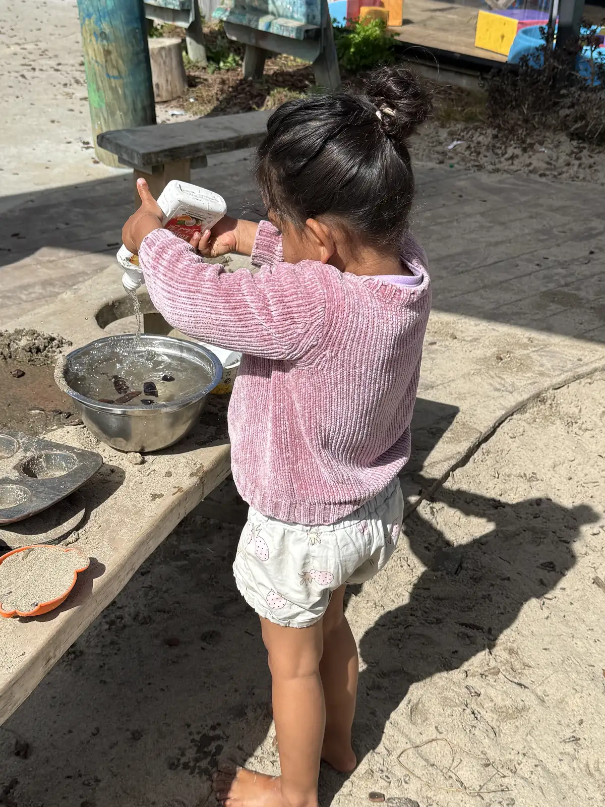 A child pouring water from a container into a metal bowl at the outdoor mud kitchen bench, sand and baking tins alongside.