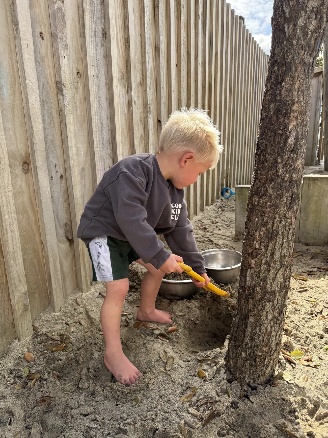A child digging a hole in the sandpit with a yellow spade, beside a tree and the timber boundary fence in the centre's outdoor area.