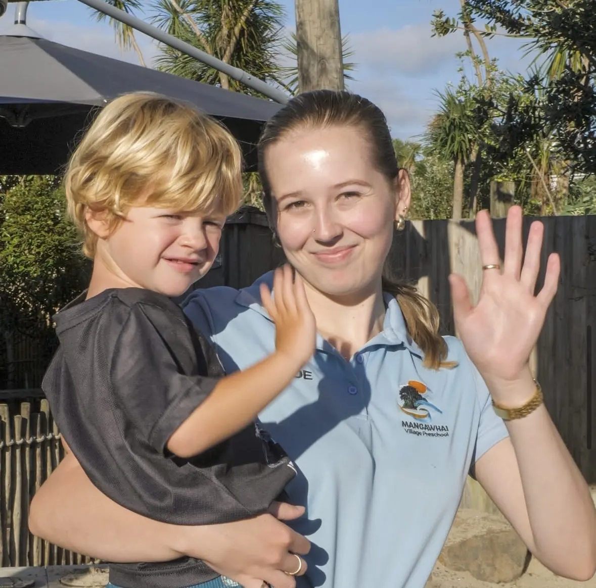 A teacher in a Mangawhai Village Preschool polo shirt carrying a child, both waving at the camera.