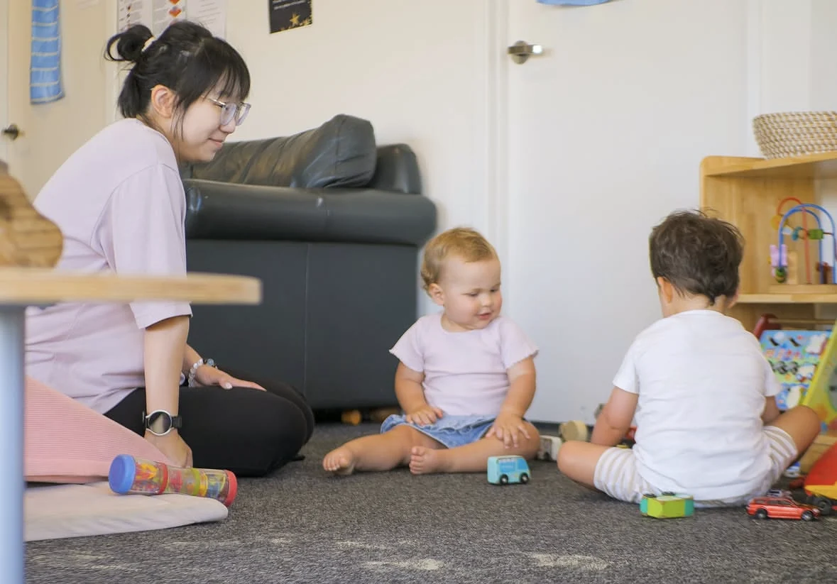 A teacher sitting on the floor of the under twos room with two babies playing with wooden cars beside her.